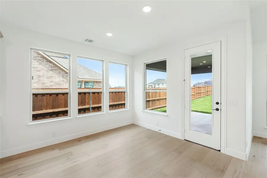 Bright empty room with light wood floors, three windows, and a glass door leading to a fenced backyard.