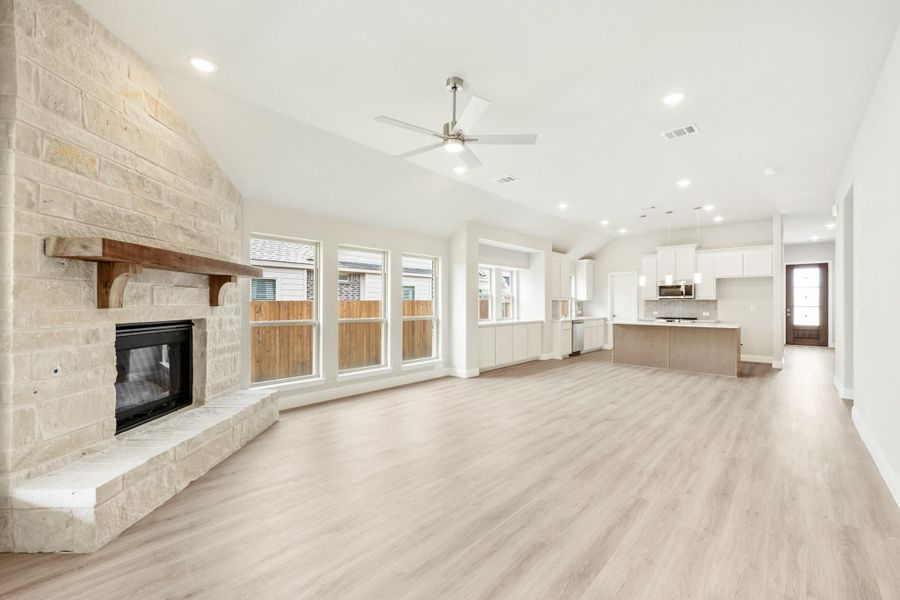 Open living area with stone fireplace, light wood floors, ceiling fan, and open kitchen in background Open living area with stone fireplace, light wood floors, ceiling fan, and open kitchen in background