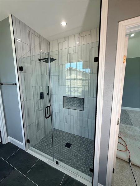 Walk-in shower featuring clear glass enclosure, gray and white patterned wall tile, black hexagon floor tile, built-in shower niche, and matte black fixtures