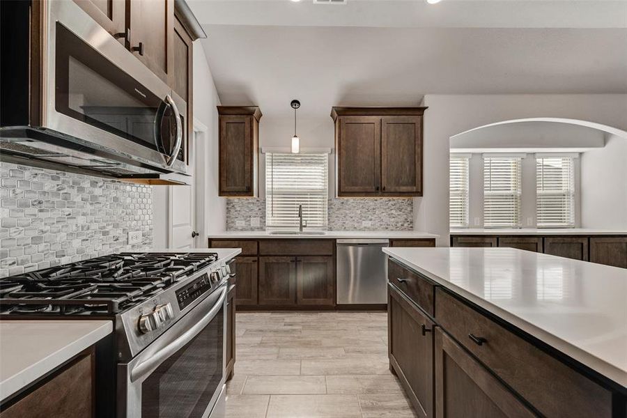 Kitchen with appliances with stainless steel finishes, dark brown cabinets, decorative light fixtures, backsplash, and light wood-style floors Kitchen with appliances with stainless steel finishes, dark brown cabinets, decorative light fixtures, backsplash, and light wood-style floors