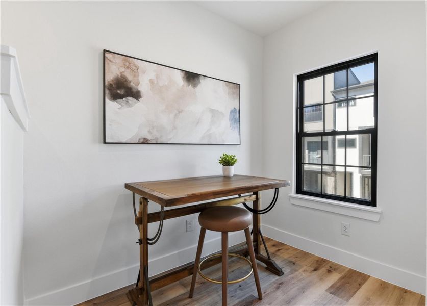 A bright, minimalist workspace featuring a wooden desk and stool, abstract wall art, and natural light from a large window.