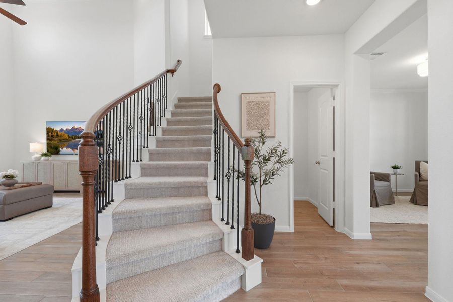 This photo features a bright, open entryway with a carpeted staircase and elegant iron railing. The space includes wood tile flooring, neutral walls, and a glimpse into a cozy living area and room. Ideal for a modern home setting.