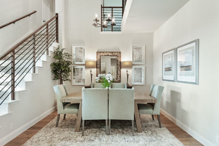 Formal dining room with wood table, six upholstered chairs, chandelier, and two-story ceiling next to staircase