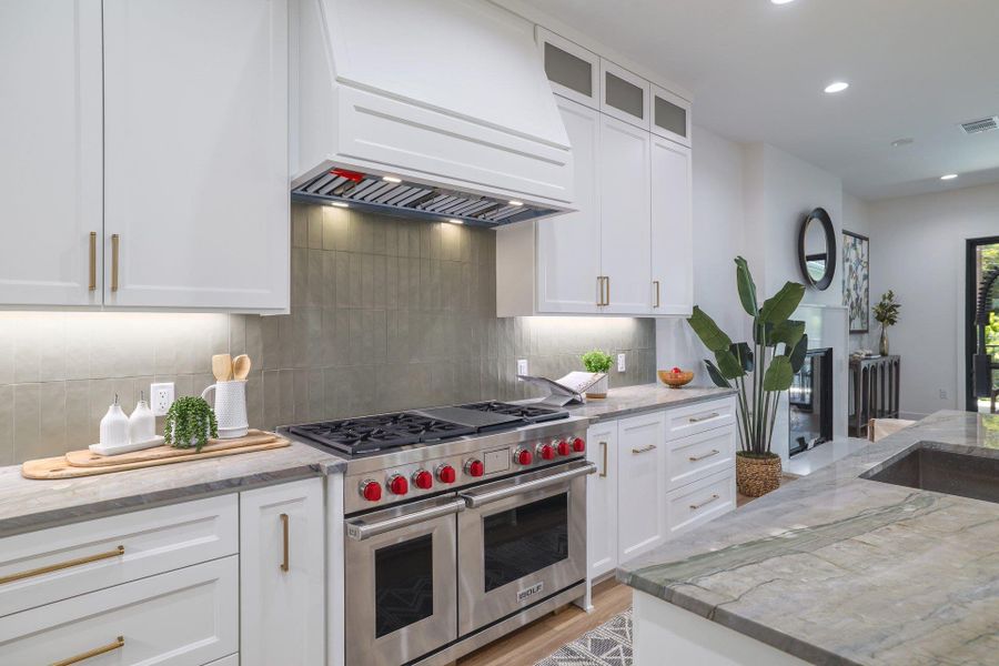 Gourmet kitchen featuring a stainless steel range with double ovens, custom vent hood, white shaker cabinetry with gold-tone hardware, textured tile backsplash, and stone countertops