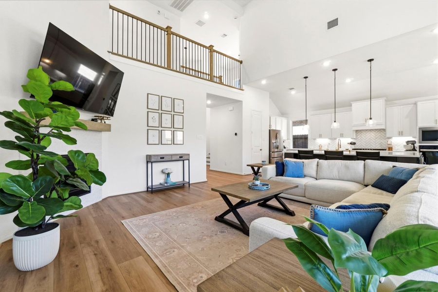 This photo showcases a spacious, open-concept living area with high ceiling, a cozy fireplace, and leads into a bright kitchen with a breakfast bar and sleek lighting fixtures. Wood look vinyl flooring and a balcony add to the airy, welcoming atmosphere.