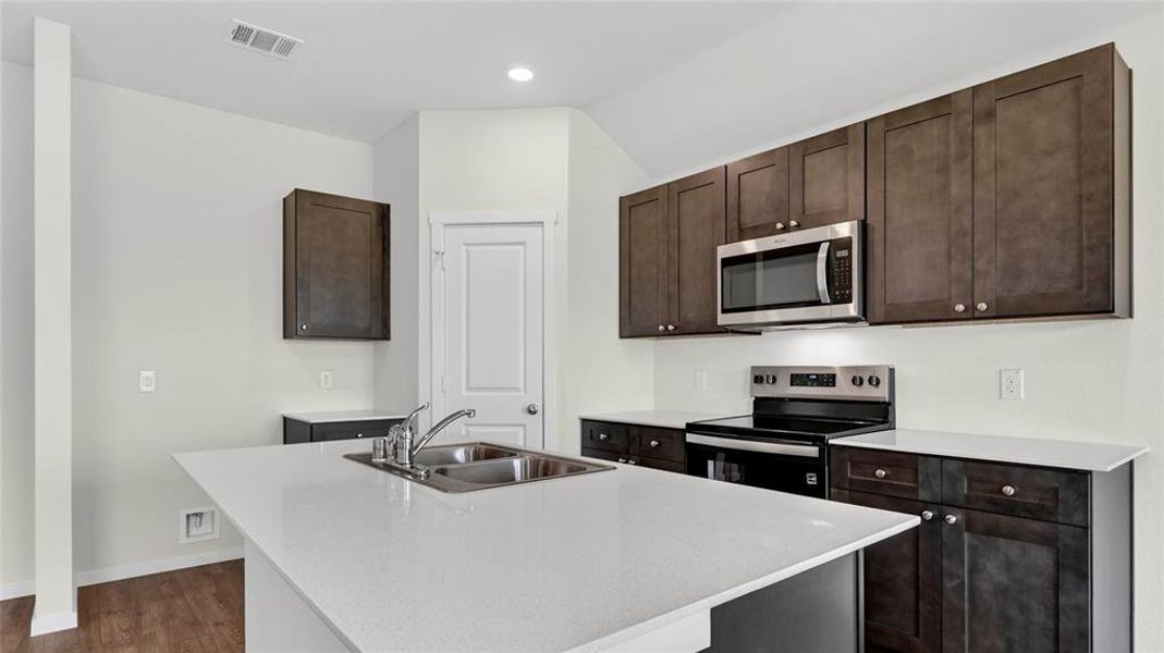 Kitchen featuring a center island with double basin sink, dark wood cabinetry, white countertops, stainless steel microwave, and wood-finish flooring
