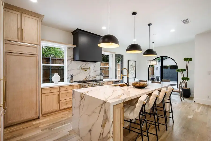 Spacious kitchen featuring a waterfall island, integrated appliances, natural wood cabinetry, white marble-look countertops and backsplash, and light wood-finish flooring Spacious kitchen featuring a waterfall island, integrated appliances, natural wood cabinetry, white marble-look countertops and backsplash, and light wood-finish flooring