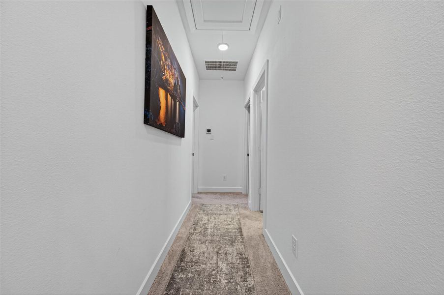 This photo shows a clean, well-lit hallway with white walls, a textured carpet, and a wall-mounted artwork. The hallway leads to multiple doors, suggesting access to various rooms. This photo shows a clean, well-lit hallway with white walls, a textured carpet, and a wall-mounted artwork. The hallway leads to multiple doors, suggesting access to various rooms.