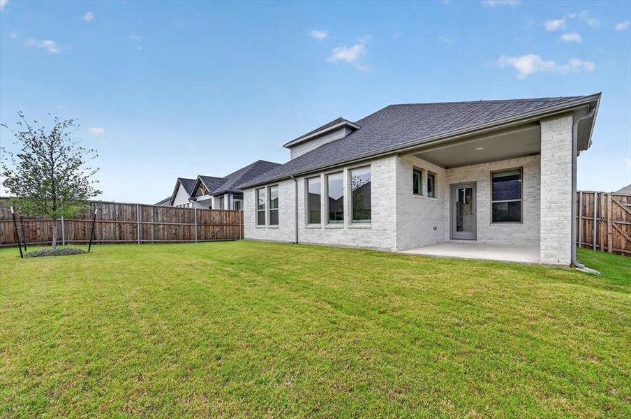 Expansive grassy backyard featuring a full privacy fence, a young tree, and a covered patio with a brick exterior