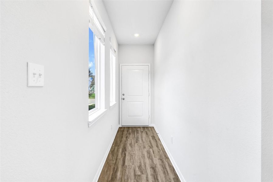 Bright hallway with clean walls and wood-style flooring. Features two large windows providing natural light and a door at the end.Please note: The image is not the actual home; it is a home with the same floor plan in the neighborhood.