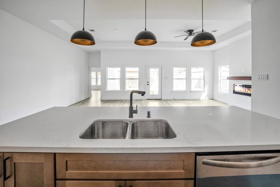 This modern open-concept living space features a sleek kitchen island with a double sink, pendant lighting, and a view of a spacious living area with large windows and a contemporary fireplace.