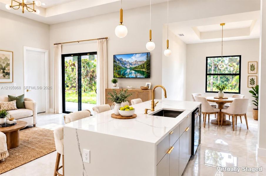 Open-concept kitchen featuring a waterfall-edge island with stone-finish countertops, brass-finish faucet, pendant lighting, recessed ceiling treatments, and large-format tile flooring
