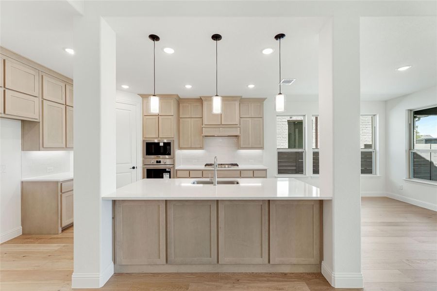 Kitchen with large island, light wood cabinets, white countertops, and three pendant lights above the sink Kitchen with large island, light wood cabinets, white countertops, and three pendant lights above the sink
