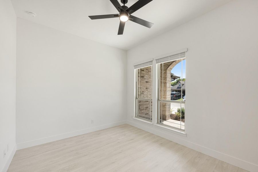 Empty bedroom with light wood floors, white walls, ceiling fan, and a window with neighborhood view