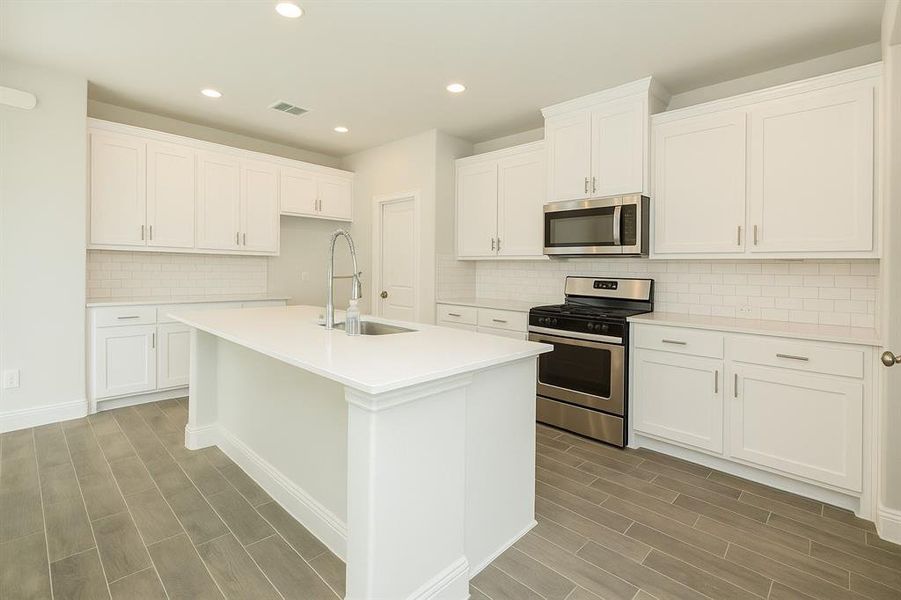 Kitchen with appliances with stainless steel finishes, backsplash, white cabinetry, wood tiled floors, and a kitchen island with sink Kitchen with appliances with stainless steel finishes, backsplash, white cabinetry, wood tiled floors, and a kitchen island with sink