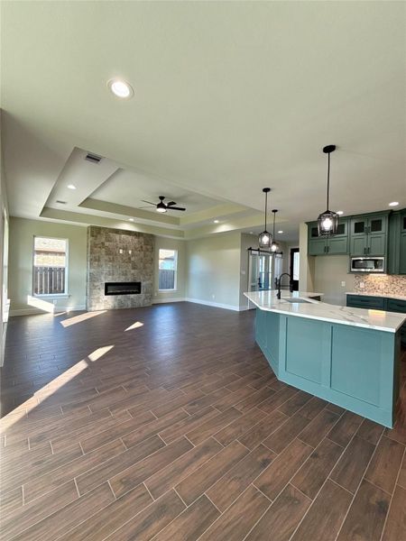 This spacious open-concept living area features elegant wood-look tile flooring, a modern kitchen with teal cabinetry and a large island, and a cozy stone fireplace. Abundant natural light enhances the room's inviting ambiance. This spacious open-concept living area features elegant wood-look tile flooring, a modern kitchen with teal cabinetry and a large island, and a cozy stone fireplace. Abundant natural light enhances the room's inviting ambiance.