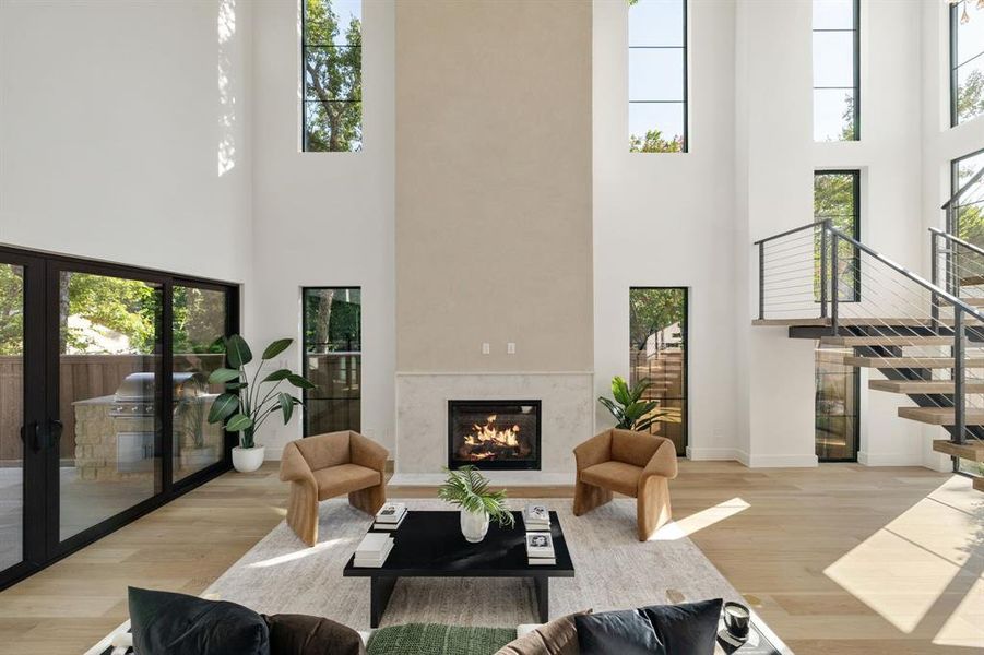Two-story living area featuring a central fireplace with stone surround, wood-finish flooring, a contemporary floating staircase with cable railings, and large sliding glass doors opening to an outdoor kitchen area