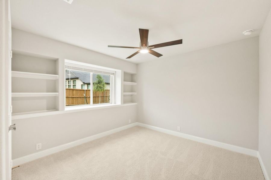 Empty bedroom with built-in shelves, ceiling fan, carpet flooring, and a window overlooking a backyard fence. Empty bedroom with built-in shelves, ceiling fan, carpet flooring, and a window overlooking a backyard fence.