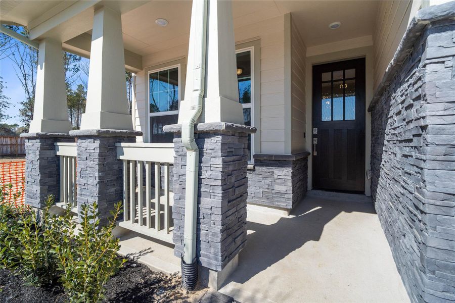 Welcome to a charming, well-maintained front porch, perfect for relaxing and enjoying the outdoors. The porch features sophisticated grey stacked stone accents around the base of the columns and the house's foundation, complemented by neutral beige siding and smooth trim.