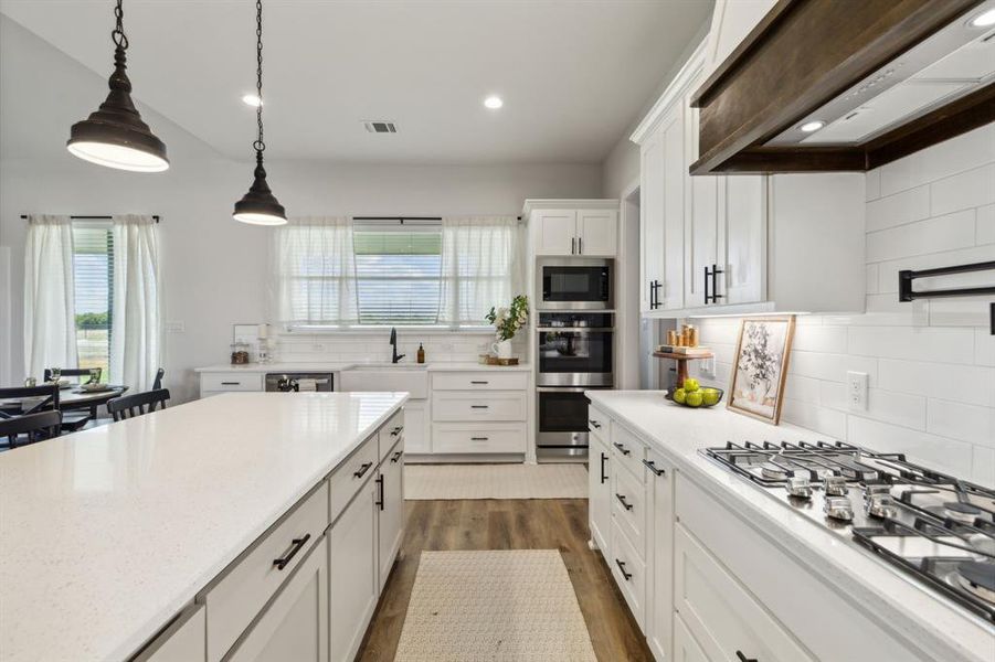 Spacious kitchen featuring a large central island with a light-toned countertop, white shaker cabinetry, stainless steel appliances, a gas range, and wood-finish flooring