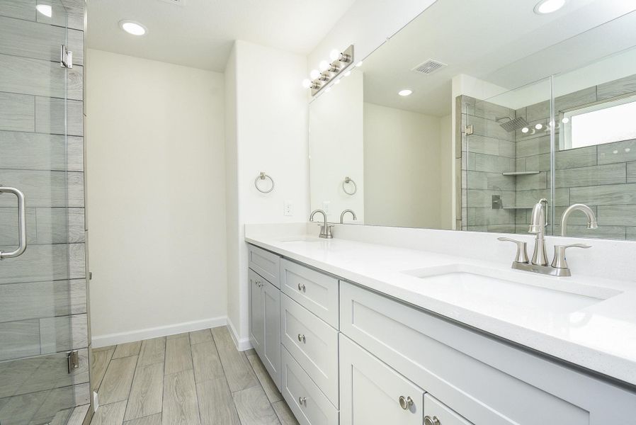 This is a modern bathroom featuring a large vanity with dual sinks, ample storage, and a spacious walk-in shower with glass doors and grey tile. The room is finished with wood-look tile flooring, offering a sleek and contemporary feel. This is a modern bathroom featuring a large vanity with dual sinks, ample storage, and a spacious walk-in shower with glass doors and grey tile. The room is finished with wood-look tile flooring, offering a sleek and contemporary feel.