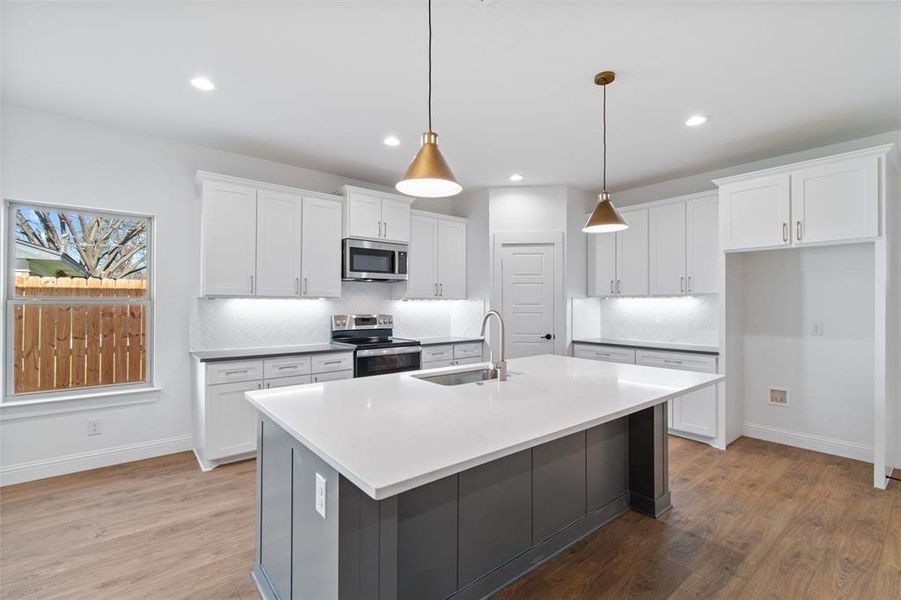 Kitchen with white cabinetry, hanging light fixtures, stainless steel appliances, tasteful backsplash, and a center island with sink