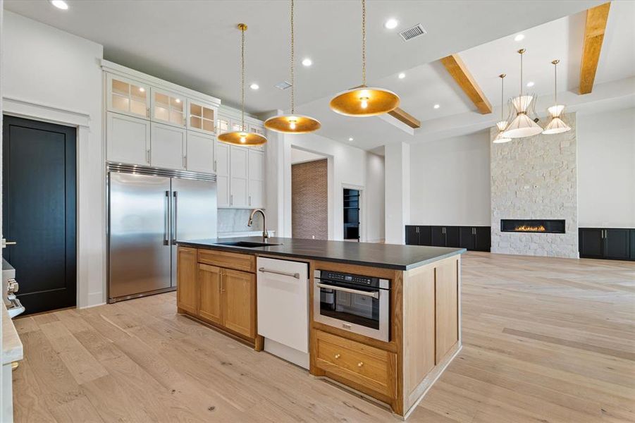 Kitchen featuring appliances with stainless steel finishes, dark countertops, a kitchen island with sink, light wood finished floors, and beam ceiling