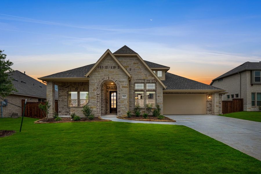 Single-story stone home with arched entry, two-car garage, and manicured front lawn at dusk Single-story stone home with arched entry, two-car garage, and manicured front lawn at dusk