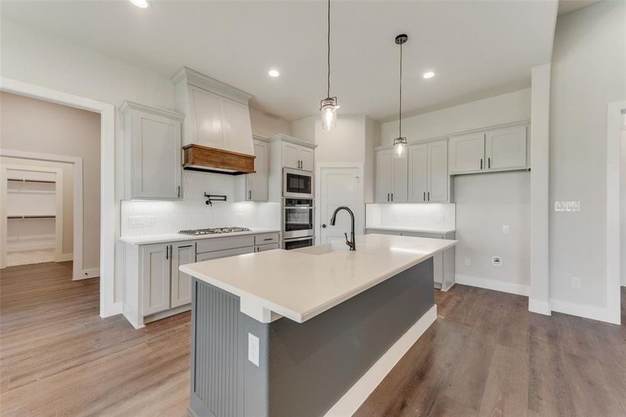 Kitchen featuring light hardwood / wood-style flooring, pendant lighting, decorative backsplash, a center island with sink, and appliances with stainless steel finishes