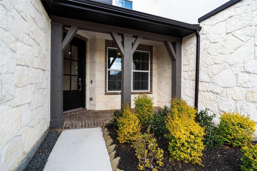 This covered front porch offers a grand first impression with its modern yet rustic aesthetic. The entrance features striking dark-stained timber beams and brackets that provide structural character against a beautiful white stone veneer façade.