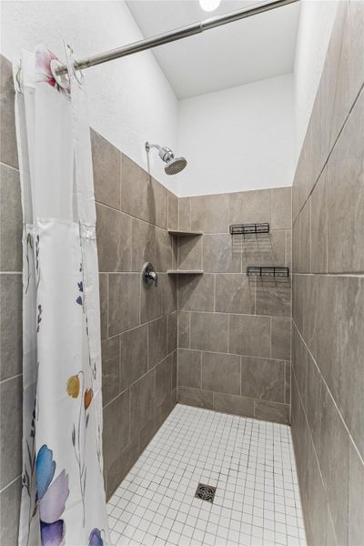 Walk-in shower featuring gray subway tile walls, white square floor tiles, a built-in corner shelf, and a wall-mounted showerhead Walk-in shower featuring gray subway tile walls, white square floor tiles, a built-in corner shelf, and a wall-mounted showerhead
