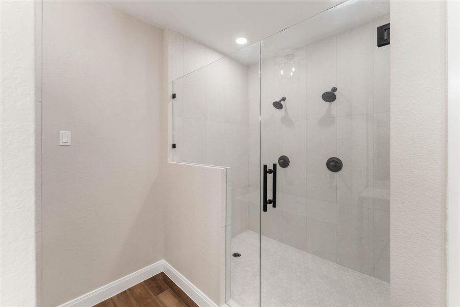 This photo showcases a modern, spacious shower with a glass door, dual showerheads, and sleek black fixtures. The neutral tile design and recessed lighting create a bright and elegant atmosphere.