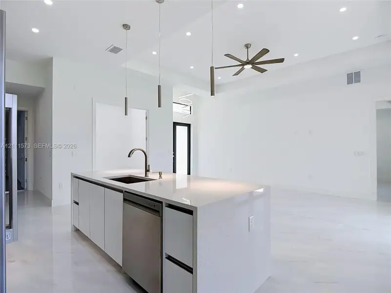 Modern kitchen island featuring a light-colored countertop, undermount sink, built-in dishwasher, white cabinetry, and a high-arch faucet, complemented by recessed ceiling lighting, three pendant lights, and a multi-blade ceiling fan