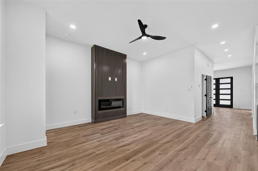 Spacious room featuring wood-finish flooring, a built-in fireplace with a fluted panel surround, recessed lighting, and a modern ceiling fan Spacious room featuring wood-finish flooring, a built-in fireplace with a fluted panel surround, recessed lighting, and a modern ceiling fan