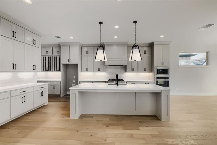 Spacious kitchen featuring light wood-finish flooring, white cabinetry with dark hardware, a large central island with a sink, white countertops, and recessed lighting Spacious kitchen featuring light wood-finish flooring, white cabinetry with dark hardware, a large central island with a sink, white countertops, and recessed lighting
