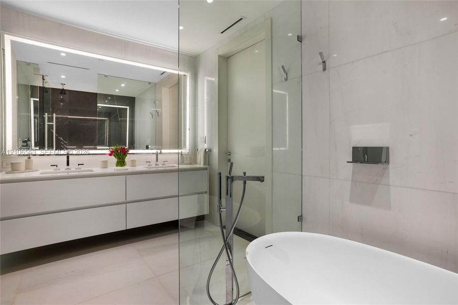 Floating vanity with dual rectangular sinks, oversized backlit mirror, floor-to-ceiling stone wall tiling, glass-enclosed shower, and freestanding soaking tub with floor-mounted faucet