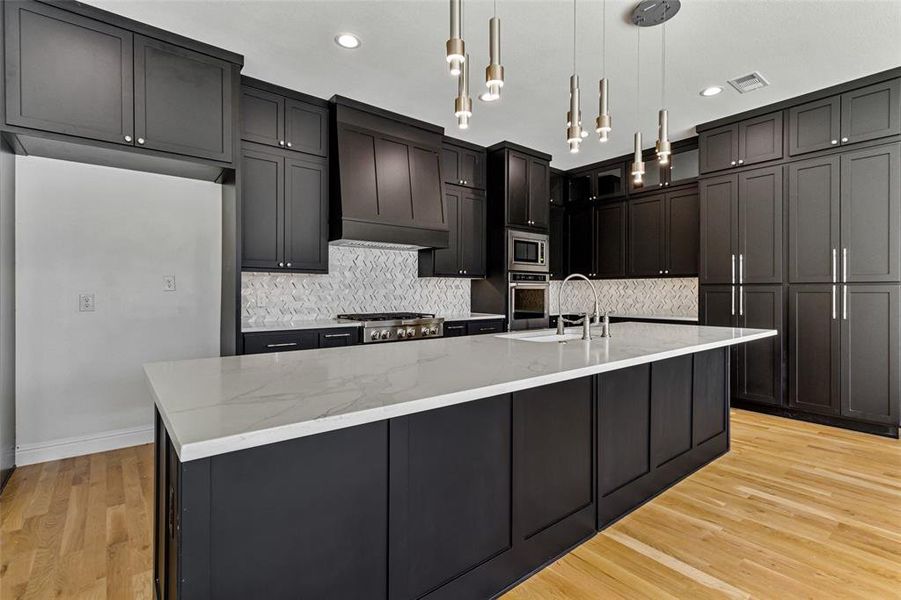 Kitchen featuring custom range hood, backsplash, stainless steel appliances, and decorative light fixtures