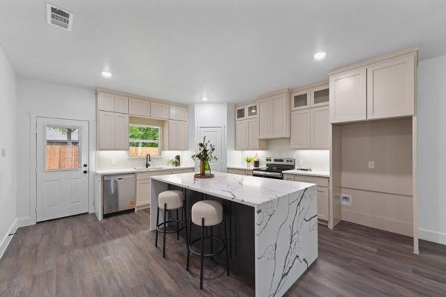 Open-concept kitchen featuring an expansive island with waterfall-edge countertop, wood-finish flooring, recessed lighting, and light-toned cabinetry with under-cabinet lighting Open-concept kitchen featuring an expansive island with waterfall-edge countertop, wood-finish flooring, recessed lighting, and light-toned cabinetry with under-cabinet lighting
