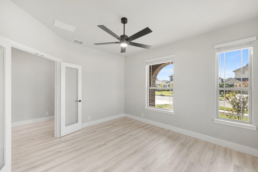 Empty bedroom with light wood floors, ceiling fan, two windows, and a glass-panel door