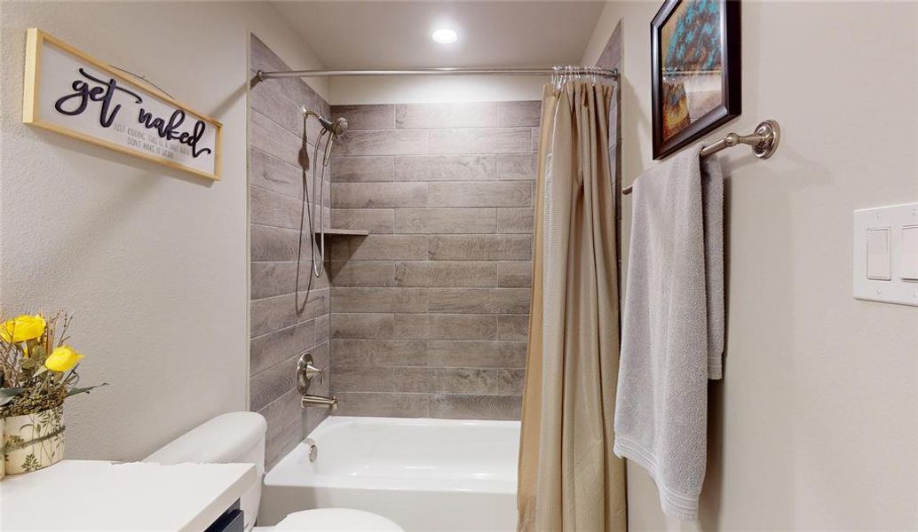 This secondary full bathroom features a modern design with a sleek white countertop, dark cabinetry, and a stylish wood-look tiled shower. Bright lighting and decorative elements add warmth and charm.