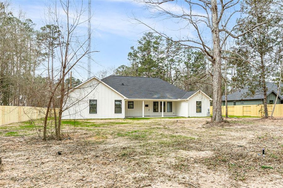 This photo shows a charming single-story home with a modern farmhouse design, featuring a white exterior and a dark roof. It is set in a spacious, partially wooded yard with a wooden fence, offering privacy and a natural setting. The large backyard provides ample space for outdoor activities.