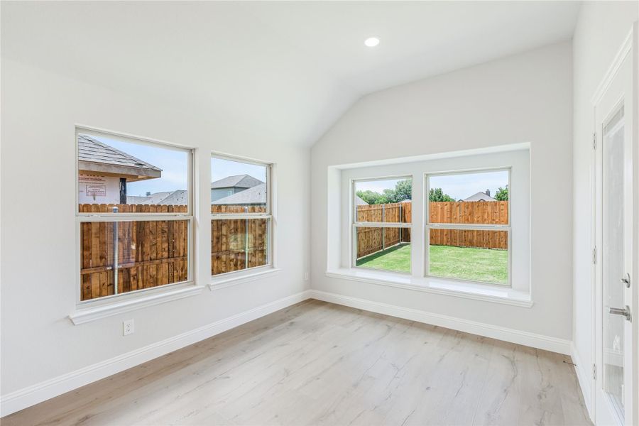 Empty room with vaulted ceiling, light wood floors, and large windows overlooking a fenced backyard