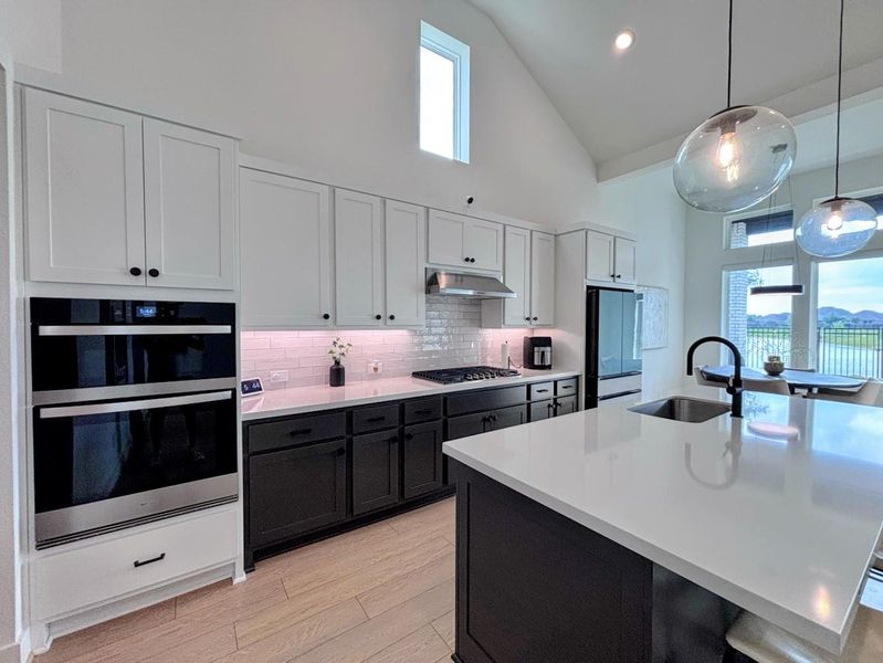 Kitchen features a bright, open design with high ceilings, sleek white cabinetry, and contrasting black lower cabinets. It includes stainless steel appliances, a large island with a sink, and pendant lighting. Natural light floods in through tall windows, offering scenic views.