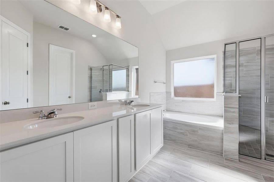 Bathroom featuring a dual vanity with white cabinetry and integrated sinks, a large wall-mounted mirror, a separate soaking tub with tile surround, a glass-enclosed shower, and light-toned tile flooring