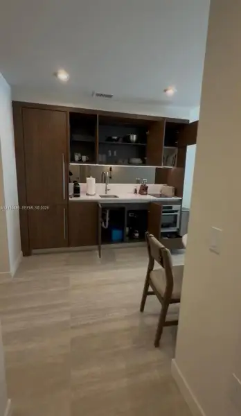 Kitchenette featuring dark wood-finish cabinetry, light-toned stone-style countertop, mirrored backsplash, integrated refrigerator, and recessed lighting