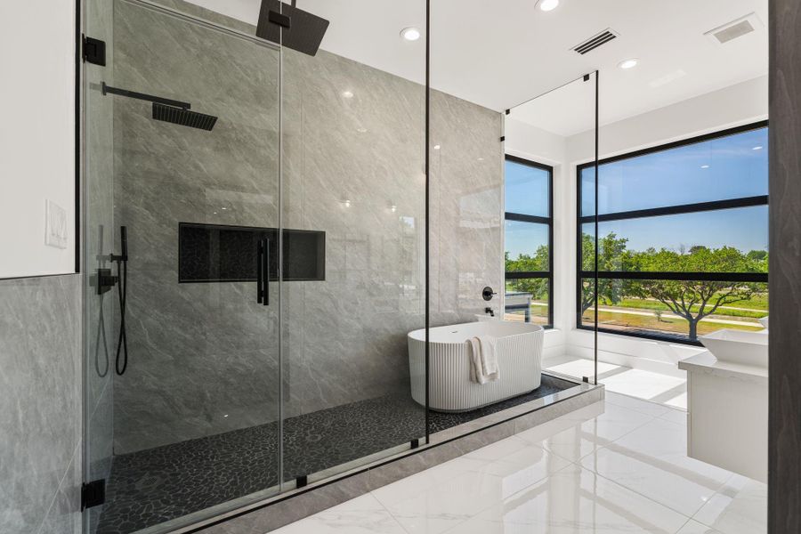 This primary bath is spa-caliber — a floor-to-ceiling large-format gray tile shower enclosed in frameless glass with a ceiling-mount rain head, matte black fixtures, and a pebble tile floor is paired with a freestanding soaking tub draped in natural light - a true everyday retreat.