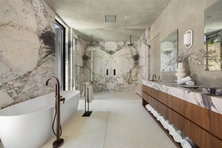 Freestanding soaking tub with floor-mounted bronze-finish faucet, full-height stone wall cladding, dual-head walk-in shower, wall-mounted floating wood-finish vanity with stone countertop, and recessed ceiling lighting