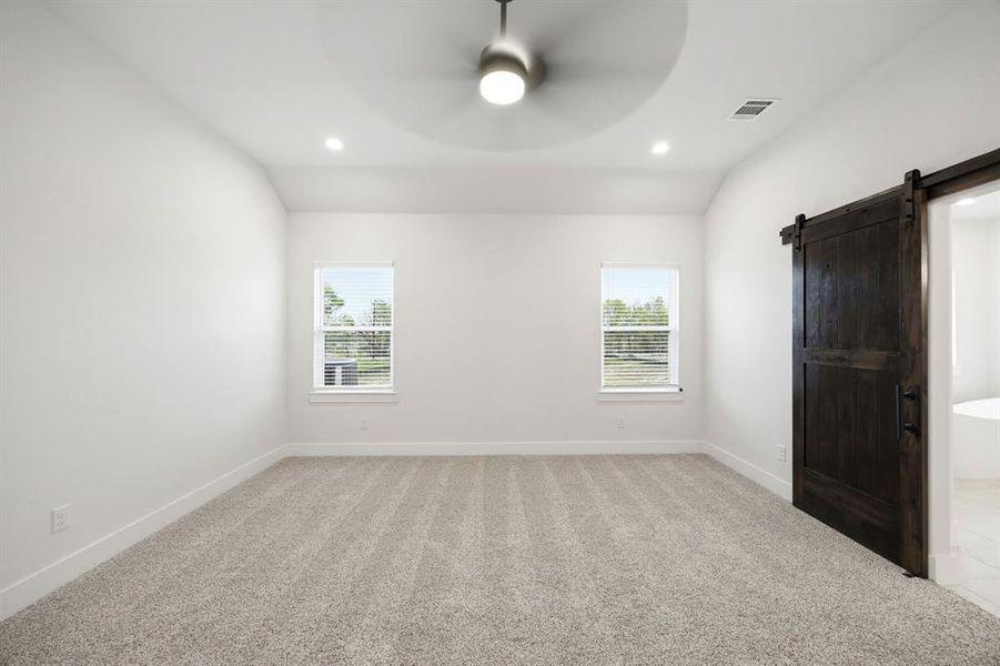 Spacious room featuring light-toned carpet flooring, two windows with blinds, a dark wood sliding barn door, and a ceiling fan with integrated lighting