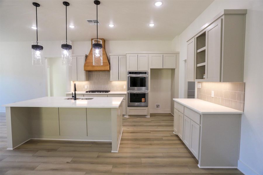 Kitchen featuring backsplash, stainless steel double oven, decorative light fixtures, and a kitchen island with sink
