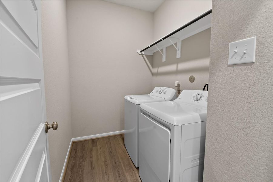 This photo shows a compact laundry room with a washer and dryer side by side. It features a simple shelf above for storage and has a light, neutral color scheme with wood-style flooring. This photo shows a compact laundry room with a washer and dryer side by side. It features a simple shelf above for storage and has a light, neutral color scheme with wood-style flooring.